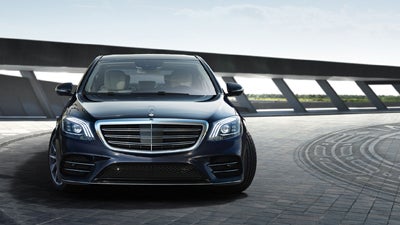 Front view of a dark blue Mercedes-Benz S-Class car. It's parked on a dark paved surface with a modern building in the background.