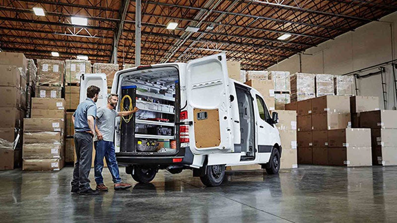 Two men next to an open white utility van with organized tools and equipment in a warehouse full of boxes.