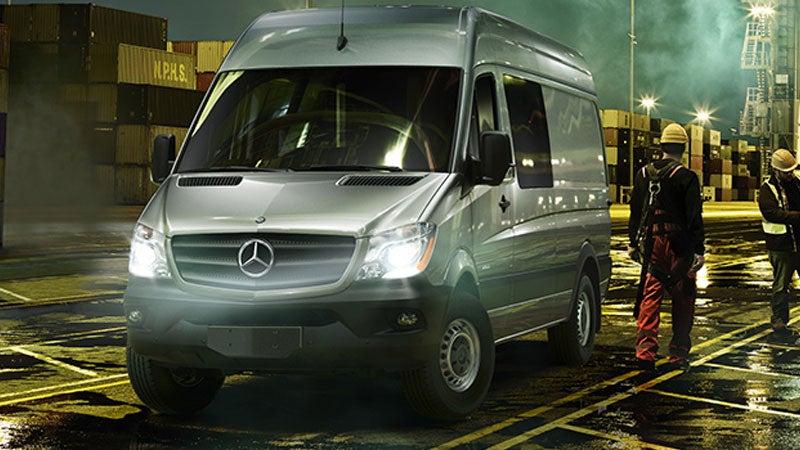 A silver Mercedes-Benz Sprinter van at a dimly lit shipping yard with two workers nearby.