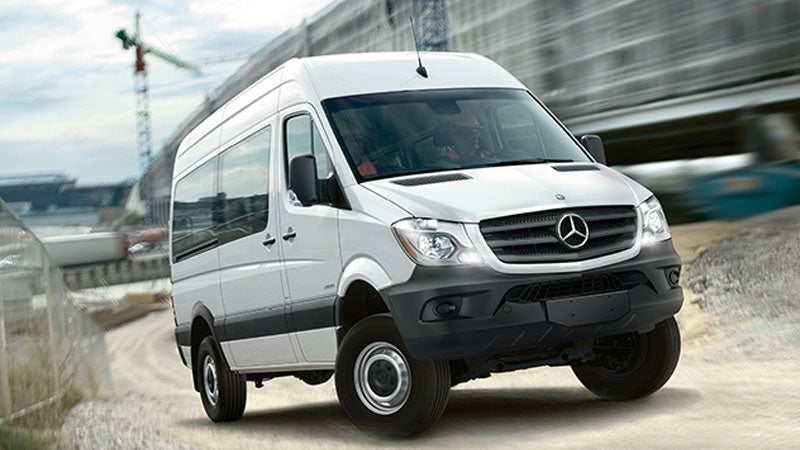 White Mercedes-Benz Sprinter van, with black bumper, driving on a dirt road next to a construction site.