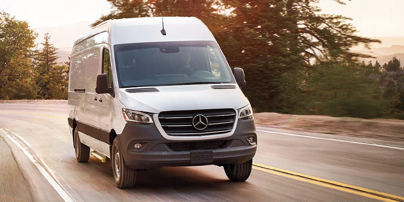 A white Mercedes-Benz Sprinter van drives on a winding road through a forest at sunset.
