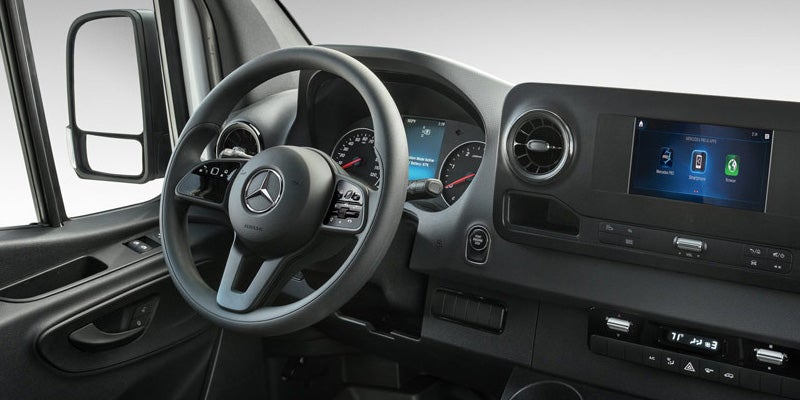 The interior of a Mercedes-Benz van: steering wheel, instrument cluster, and infotainment system showing app icons.
