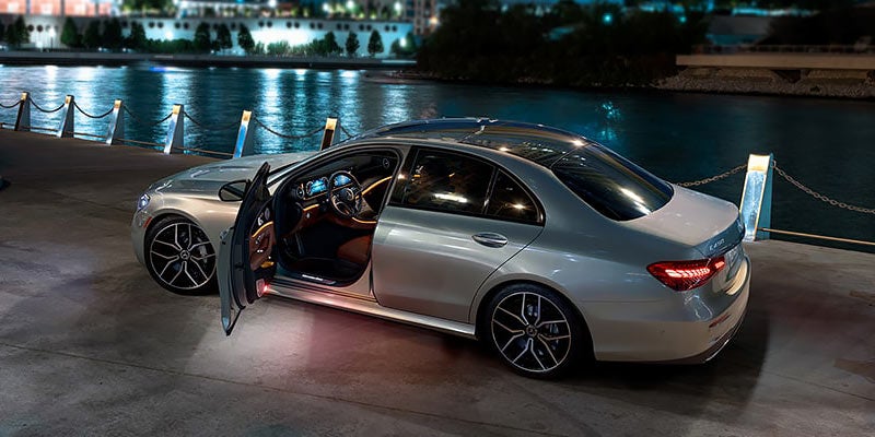 Silver Mercedes-Benz E-Class sedan at night, parked near a body of water, doors open, interior lights on.
