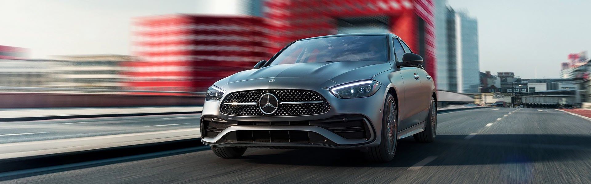 A gray Mercedes-Benz C-Class sedan drives on a blurred urban road, with motion blur indicating speed.