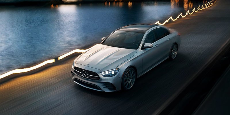 A silver Mercedes sedan driving on a dark highway next to a river with city lights reflecting on the water.