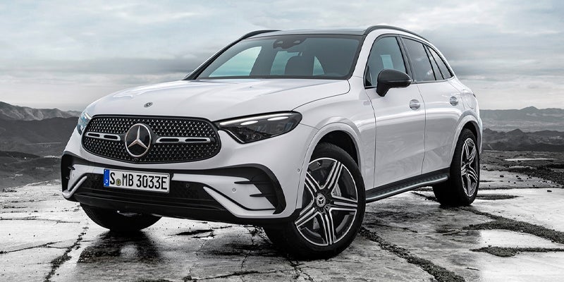 White Mercedes-Benz GLC SUV parked on a rough, stony surface with mountains and a cloudy sky in the background.