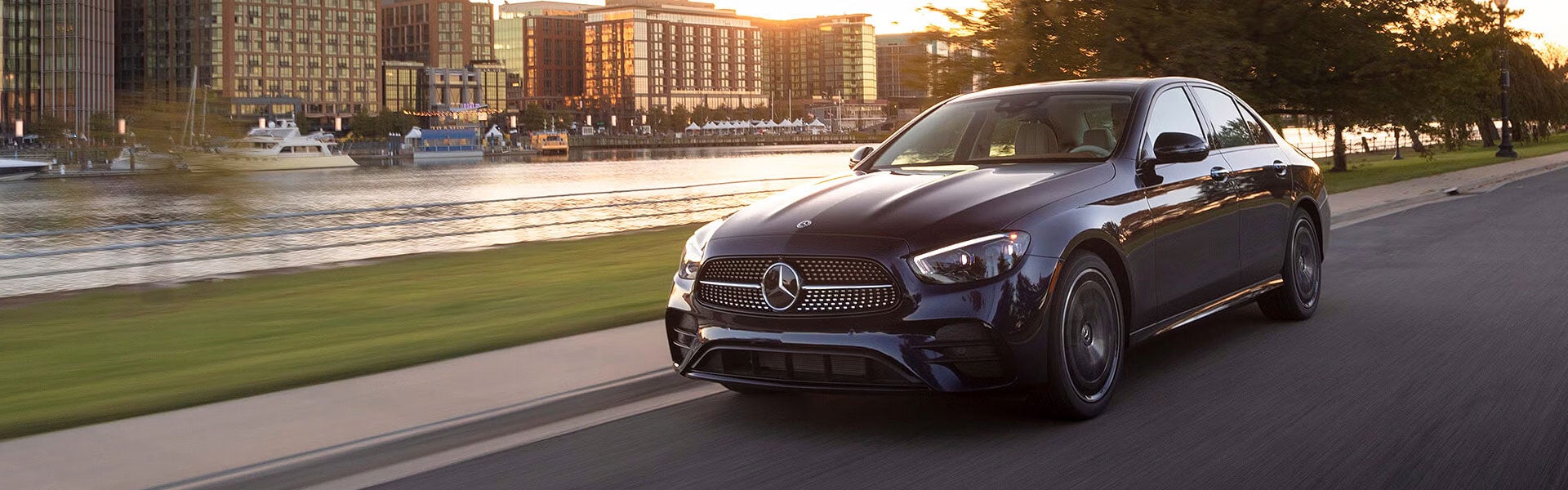 A dark Mercedes-Benz sedan driving along a waterfront city road at sunset