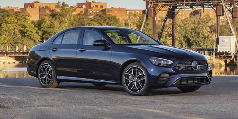 A dark blue Mercedes-Benz E-Class sedan is parked on asphalt with a water feature, trees, and buildings in the background.