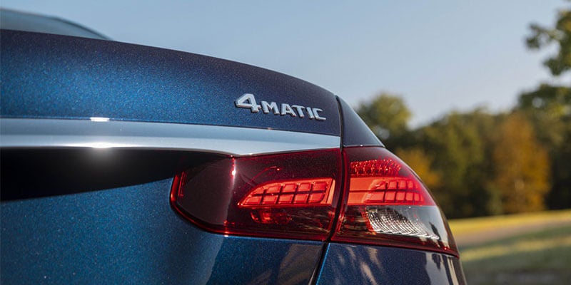 Rear view of a dark blue car, showing the 4MATIC emblem and a red taillight under a blue sky.