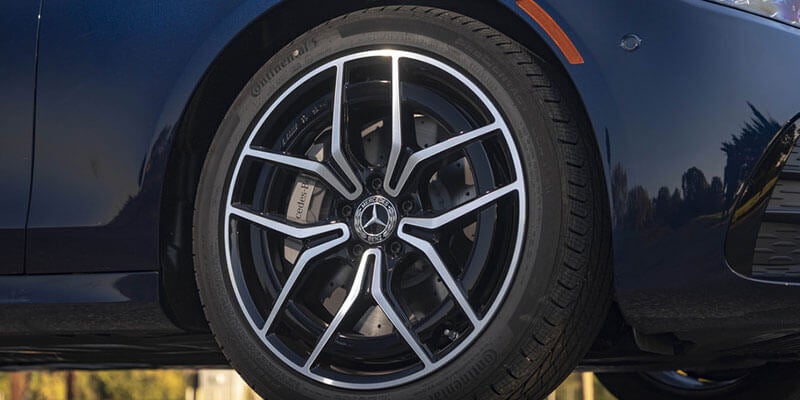 A close-up of a glossy, dark metallic blue car's passenger side front wheel and fender. The wheel has a black rim with silver spokes, a Mercedes-Benz logo, and a visible brake caliper.