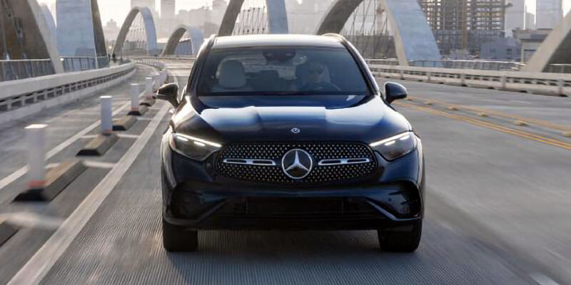 Front view of a dark Mercedes-Benz GLC SUV driving on a bridge with arched supports, distant city buildings visible.