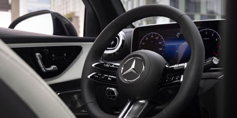 Close-up of a blurred black leather steering wheel with the Mercedes-Benz logo, dashboard screen, and car door controls.