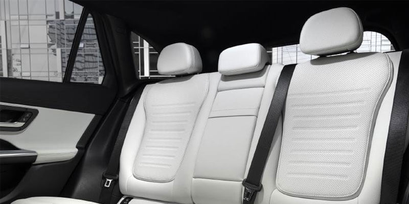 Inside a car showing white leather rear seats with headrests and black seatbelts, with a window view of city buildings.