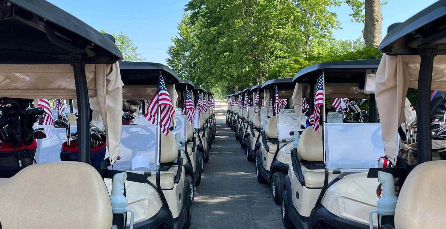 Two rows of golf carts, each with small American flags, parked on a paved path lined with green trees.