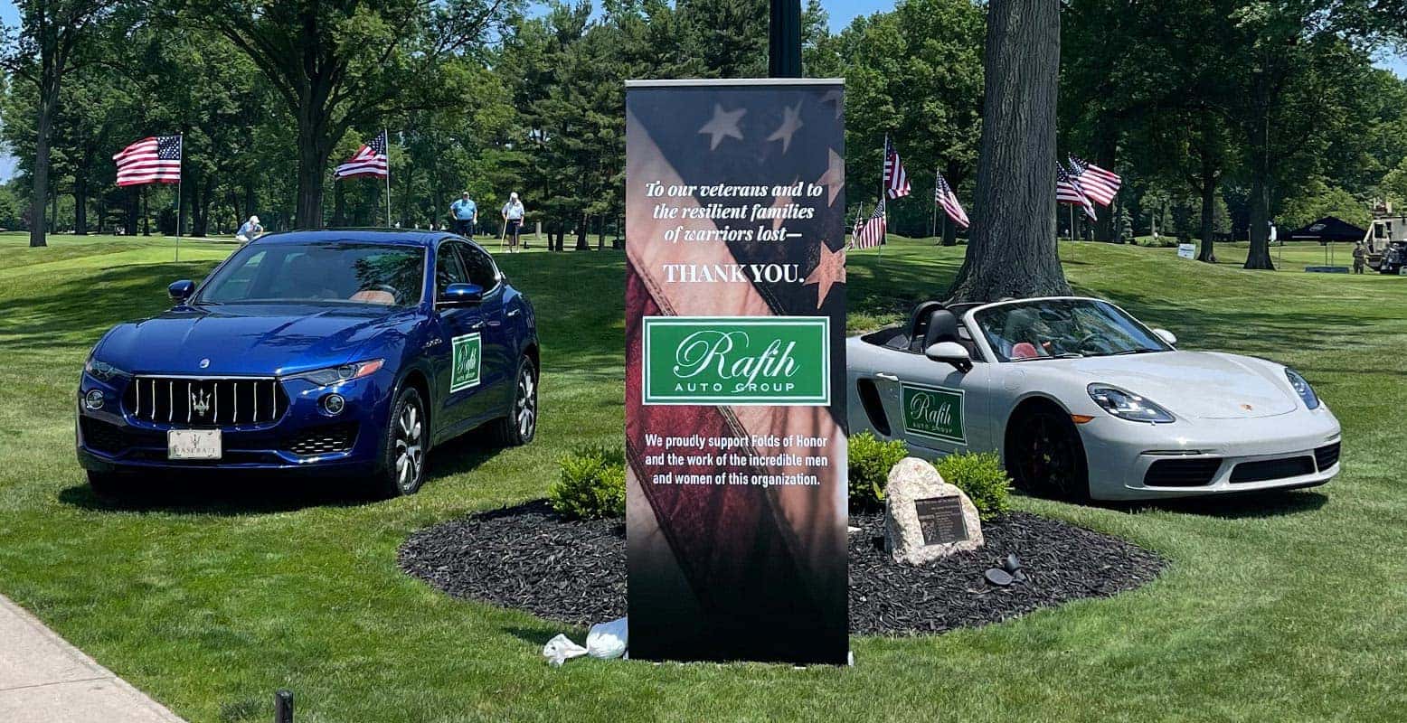 Two luxury cars in a grassy area with a banner thanking veterans and their families from Rafih Auto Group.