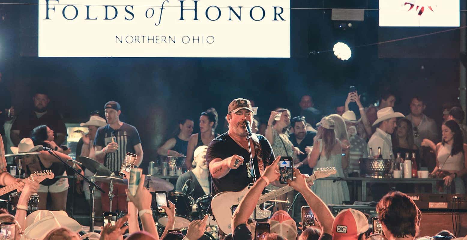 A singer performs live on stage, wearing a cap and black shirt, addressing the audience. Many people record with phones.