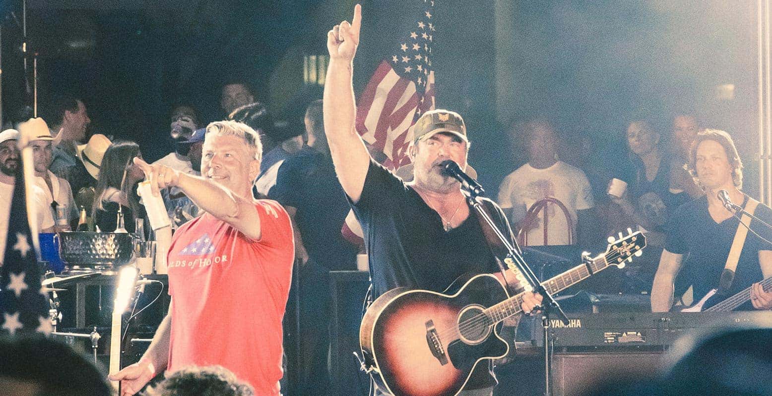 A man in a black shirt and hat sings into a mic while playing guitar, arm raised. Another man in red points.