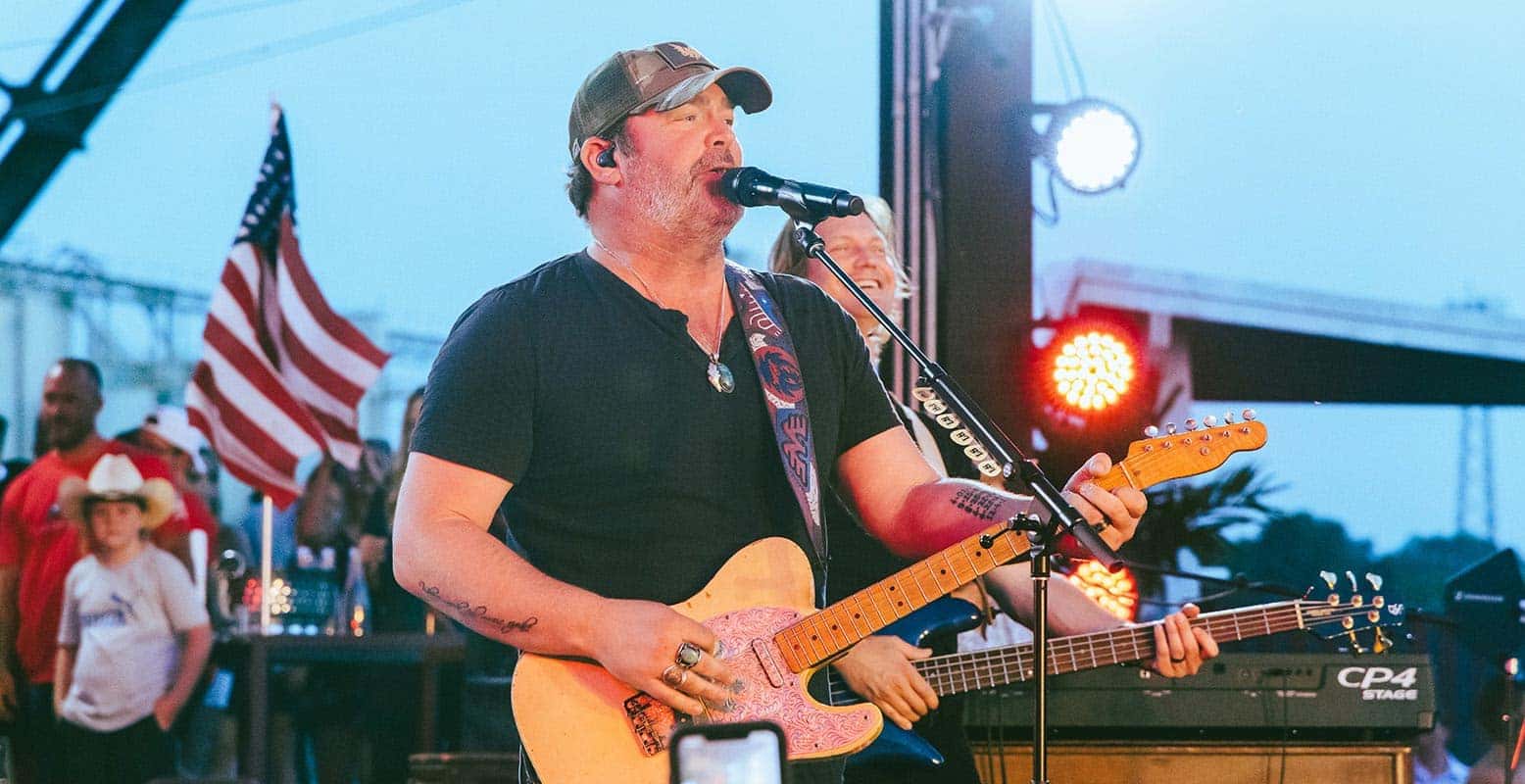 A man in a baseball cap sings into a microphone, playing a patterned guitar on stage, with an American flag behind him.
