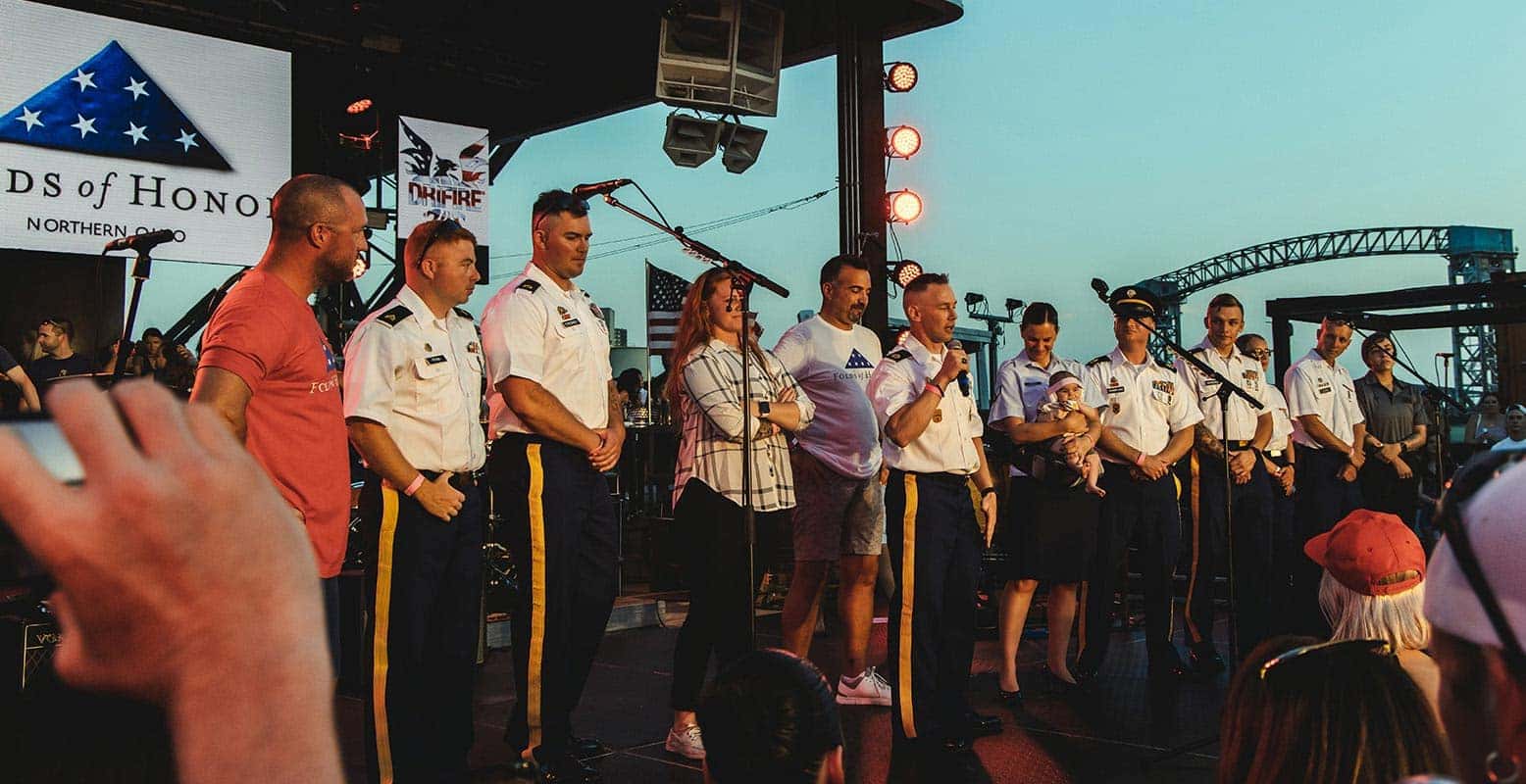 A man in a backwards cap and sunglasses points forward, among a crowd with a US flag and a large screen.