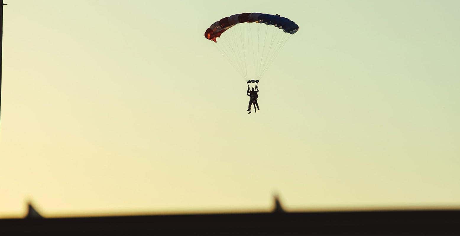 Two skydivers descend with an open parachute against a pale yellow sky.
