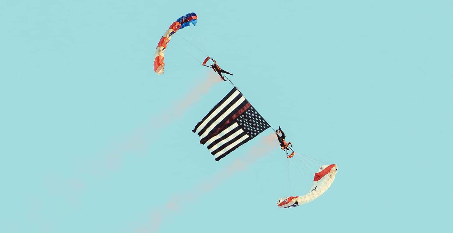 Two parachutists descend with an American flag under a light blue sky. One carries the top while the other carries the bottom.