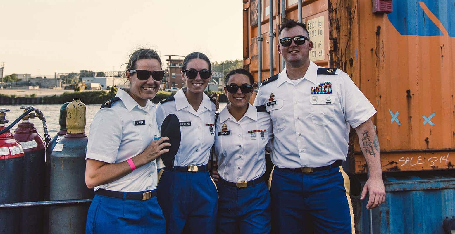 Four individuals in military uniform and sunglasses stand together smiling. They are on a boat with gas tanks visible.