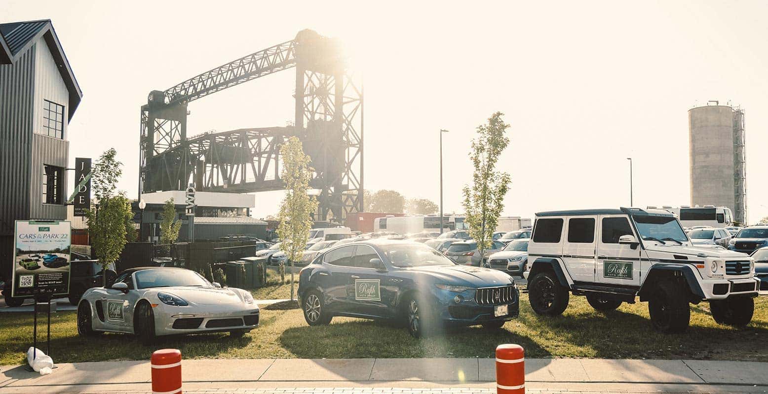 Three luxury cars, a Porsche, Maserati, and Mercedes-Benz, are parked on grass with a bridge in the background.