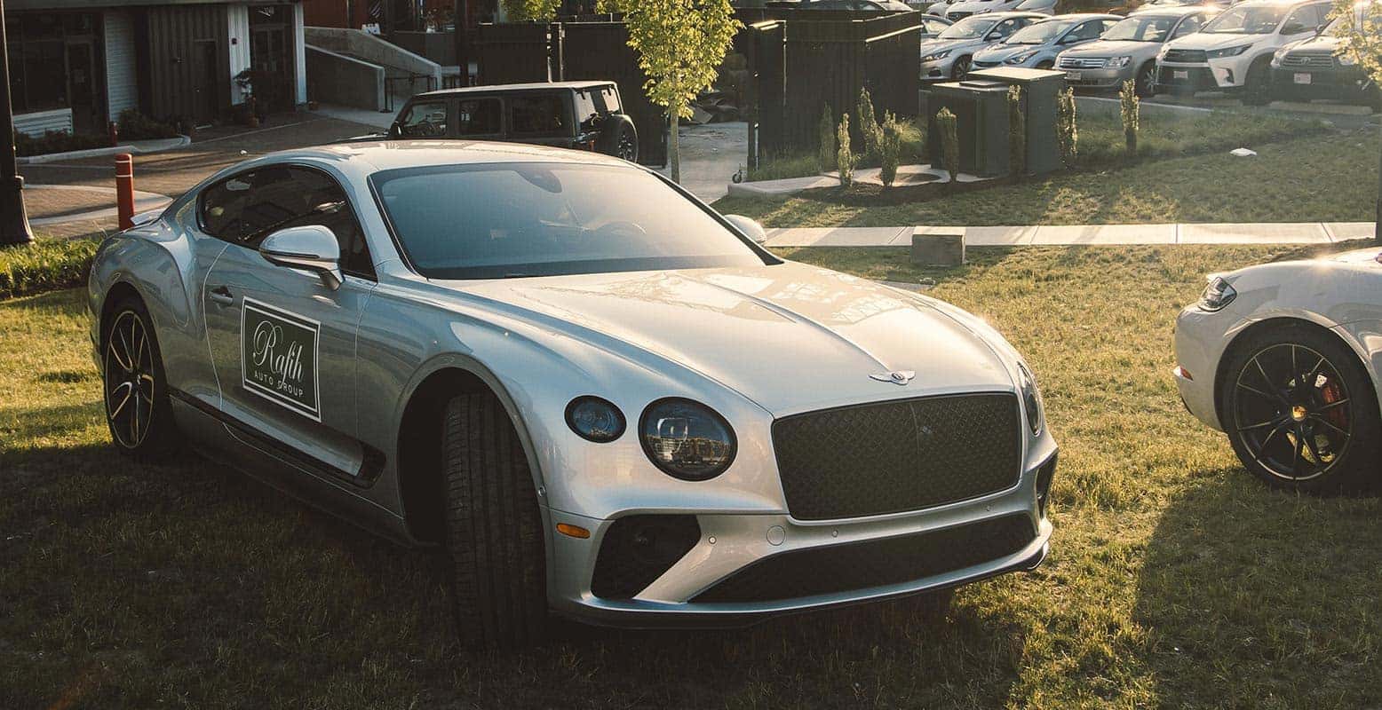 Silver Bentley Continental GT parked on grass, with a Rafith Auto Group logo on the door. Another car's front tire is visible to the right.
