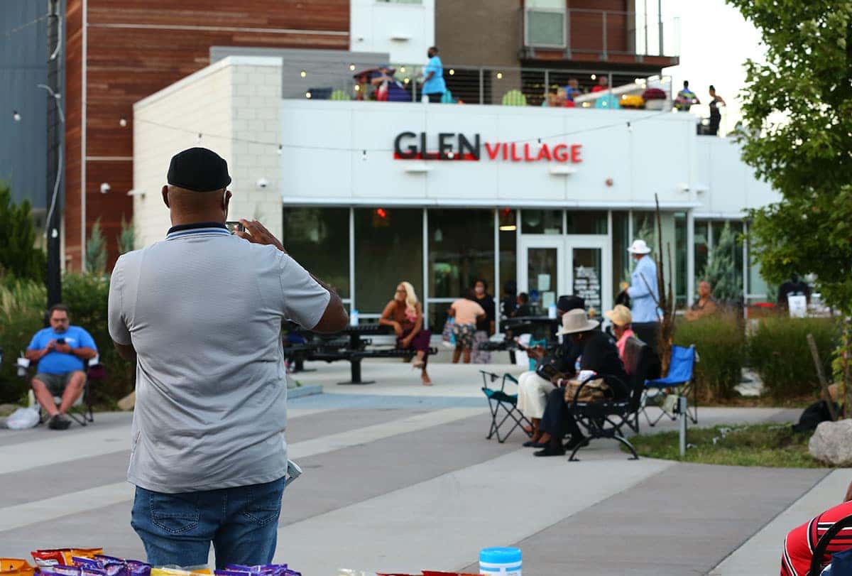 A woman in a white dress and jean jacket smiles next to a blue sports car with 