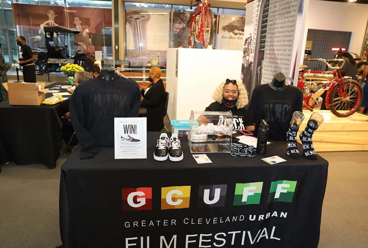 A person in a gray shirt and black cap takes a photo in front of a building with a GLENVILLAGE sign