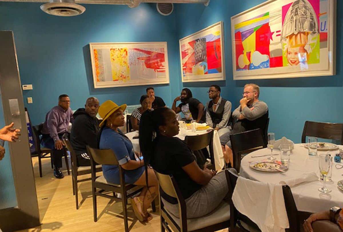 Diverse group of people seated around tables in a room with blue walls and colorful framed art.