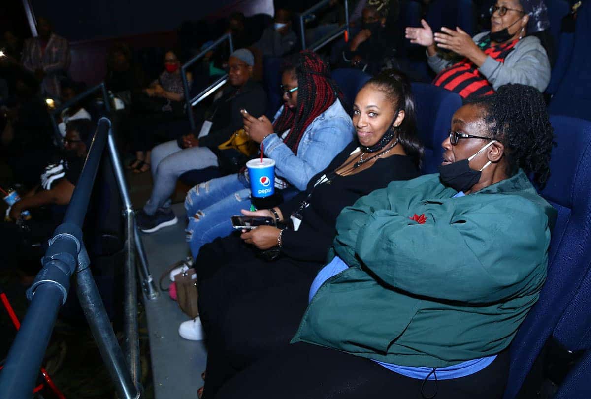 A group of people sitting in a movie theater, some looking forward, others on their phones. Most are wearing masks.