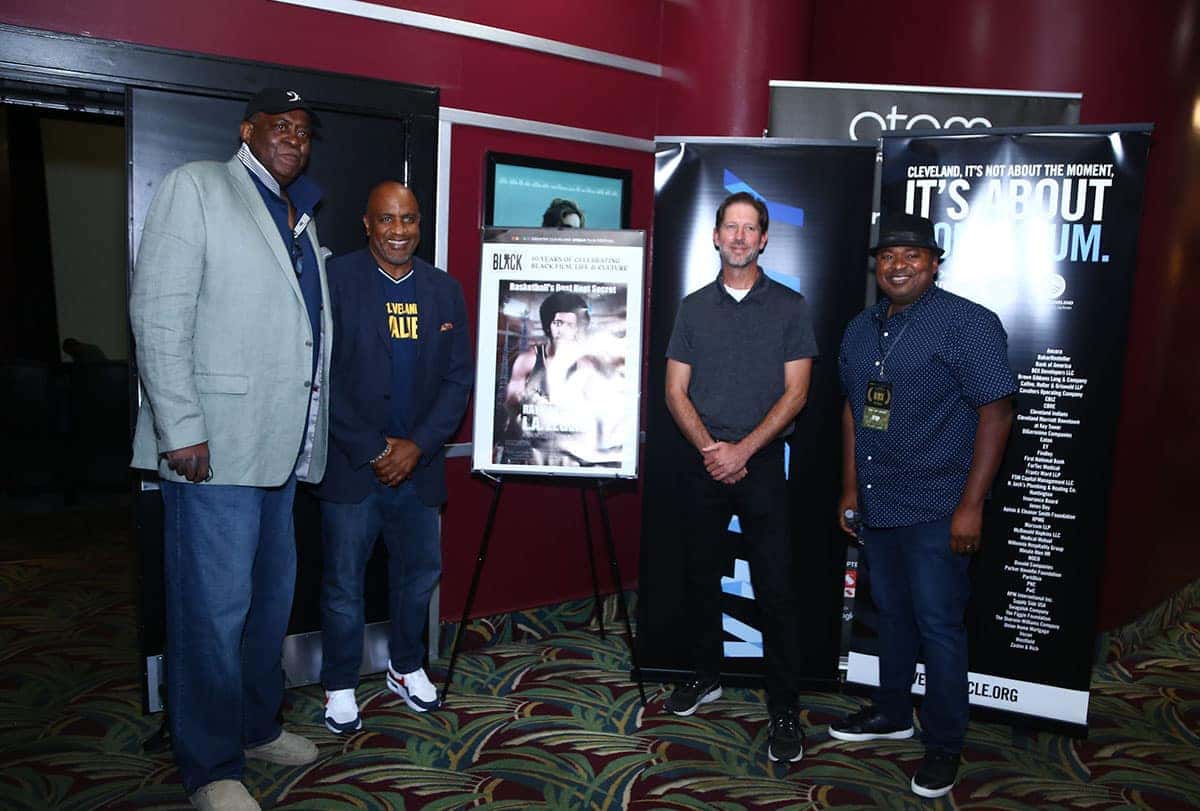 Four men stand in front of black banners at an event. A poster in the middle features a man boxing.