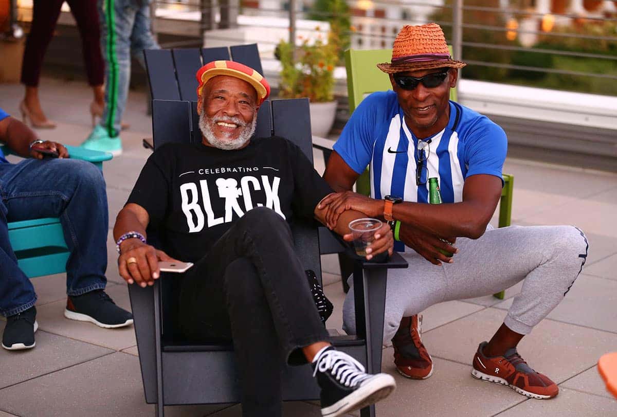 Two Black men relaxing outdoors—one in an armchair with a red-yellow hat, the other crouching in a straw hat and striped shirt.