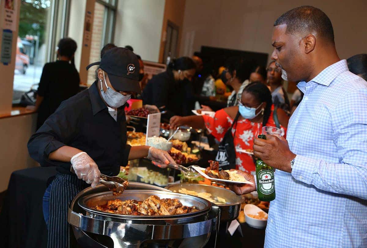 A caterer wearing a mask and gloves serves food from a chafing dish to a man holding a beer bottle and a plate.