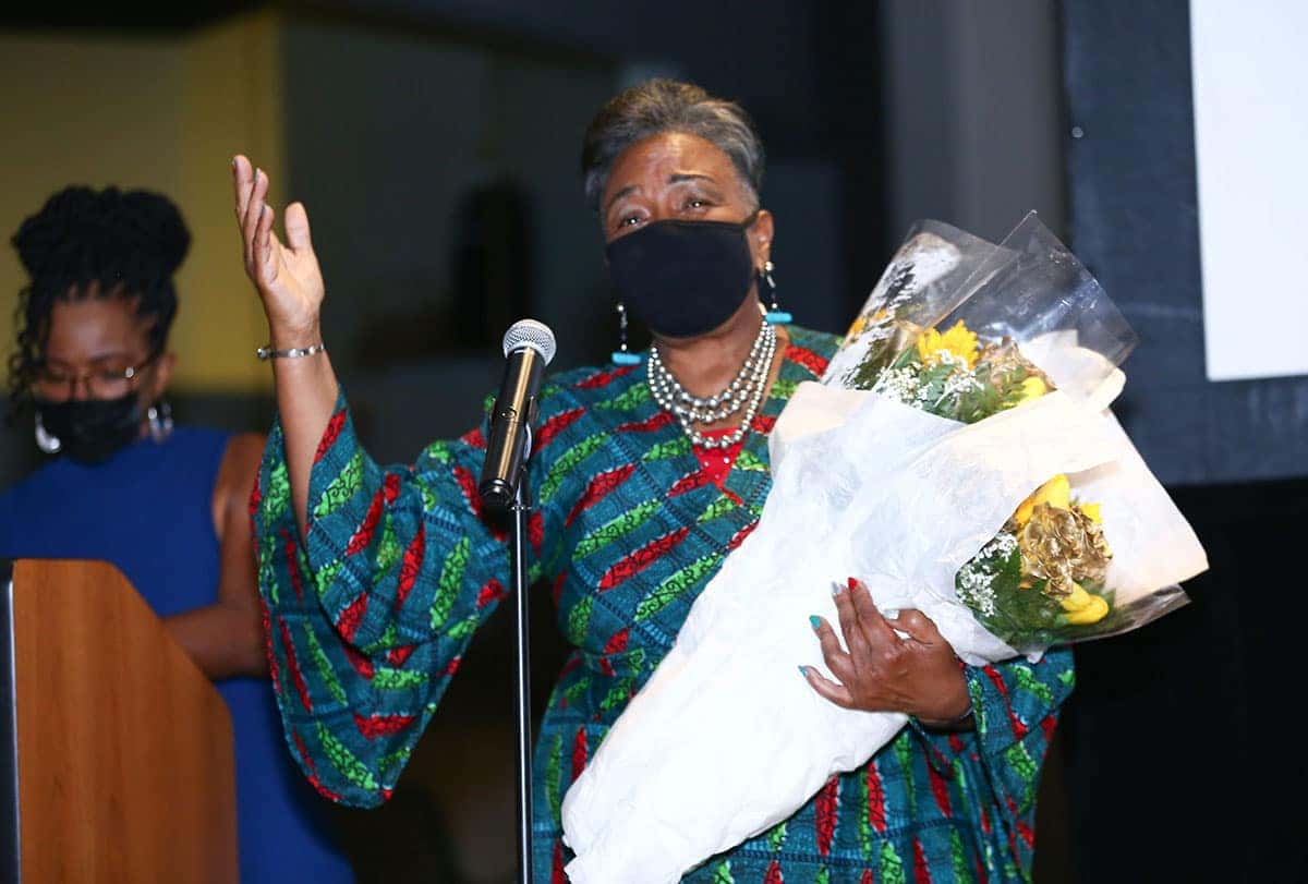 A woman in a colorful dress speaks at a microphone, holding bouquets of flowers. A second woman stands behind her.