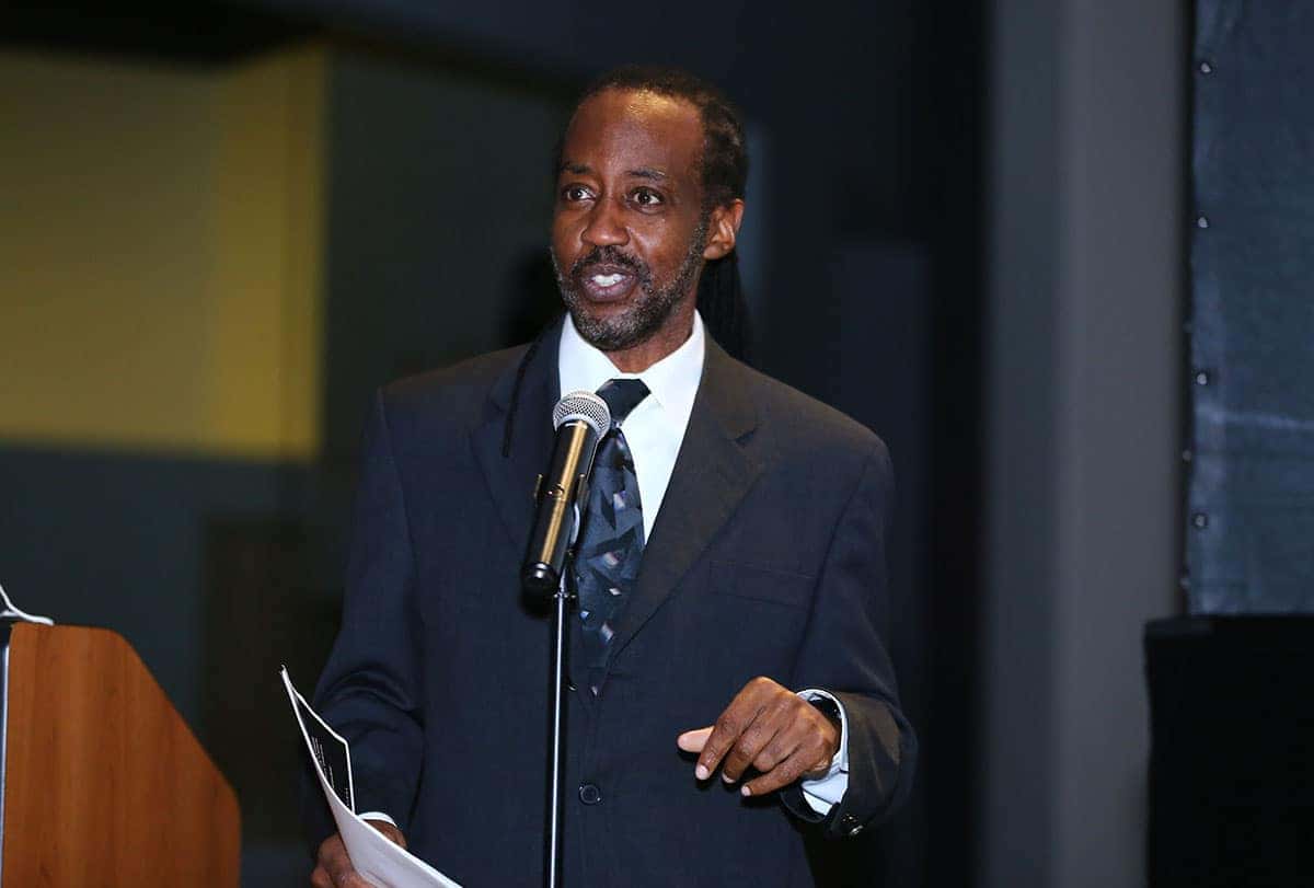 A Black man in a dark suit speaks into a microphone, holding papers in his left hand, against a dark, blurred background.