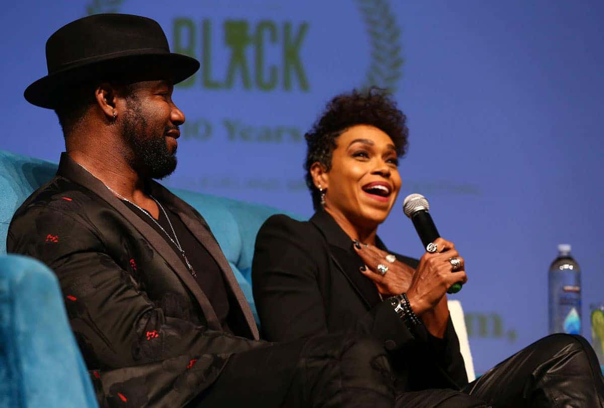 Two people at a conference. A man in a fedora smiles while looking right. A woman with a microphone laughs.