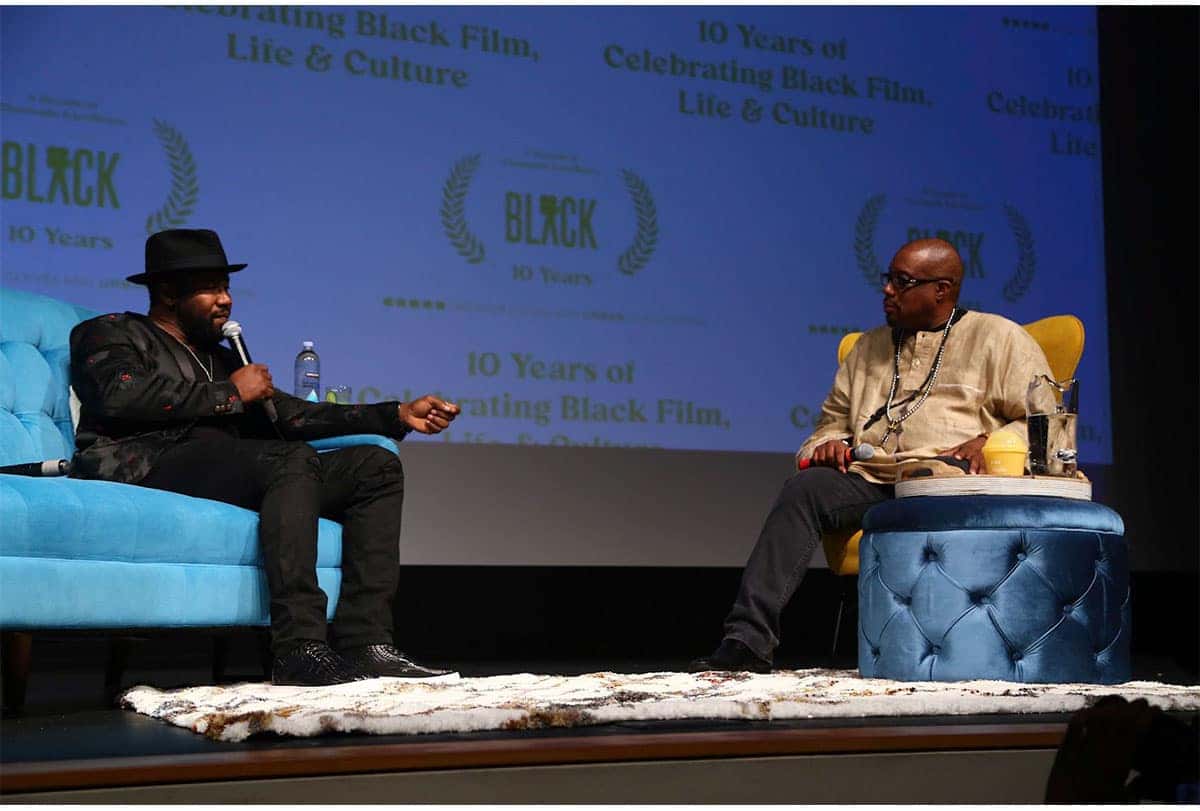Two men in chairs on a stage with a screen behind them with 10 Years of Celebrating Black Film, Life & Culture written on it.