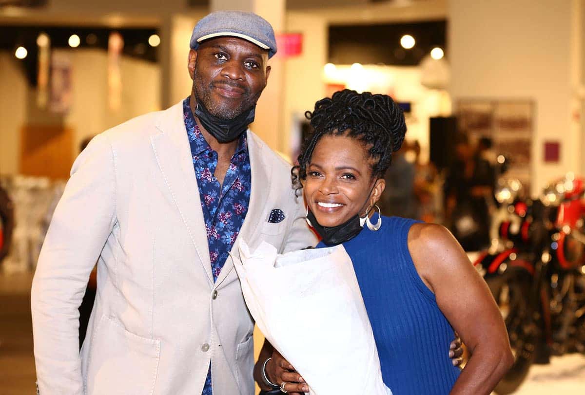 A smiling man in a tan jacket, blue patterned shirt, and gray cap stands next to a smiling woman in a blue top. Both wear face masks around their necks.