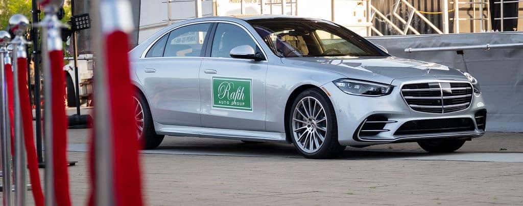Silver Mercedes-Benz sedan with a Rafih Auto Group decal on the side, parked beside a red-roped stanchion.