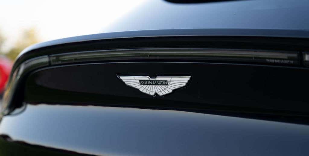 A close-up of the Aston Martin logo, a silver winged emblem with ASTON MARTIN across the center, on a black car.