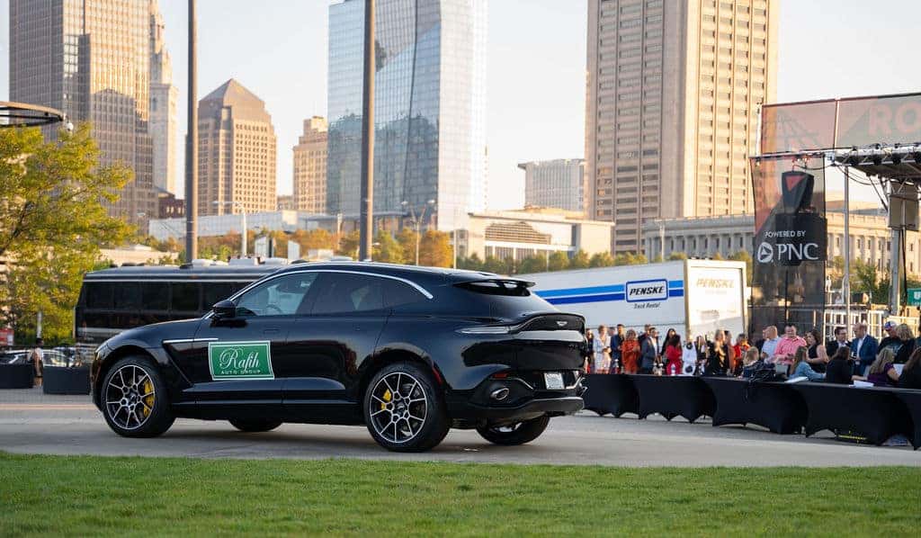 A black luxury SUV with Rafih Auto Group on the side is parked outside, city skyline in background.