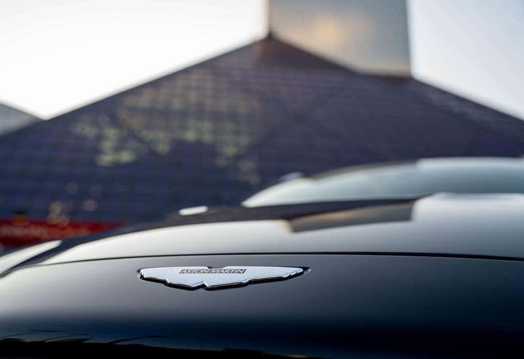 An Aston Martin car's hood with its winged logo, reflecting a blurred, angular building in the background.
