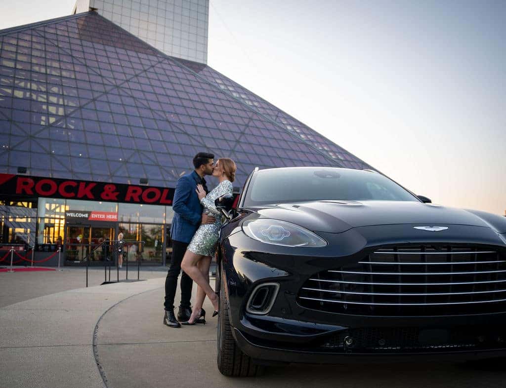 A man and woman kiss leaning against a black Aston Martin SUV in front of the Rock & Roll Hall of Fame.