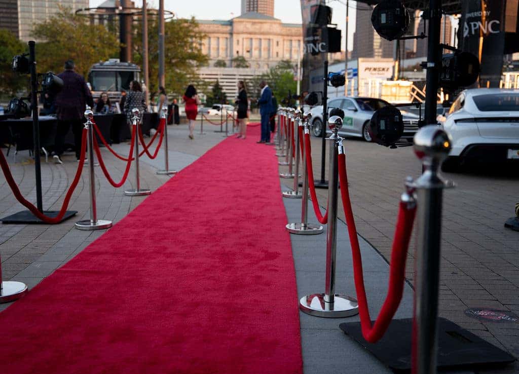 A long, red carpet stretches down a paved street. Silver stanchions with red velvet ropes line both sides. People and cars are visible in the background.