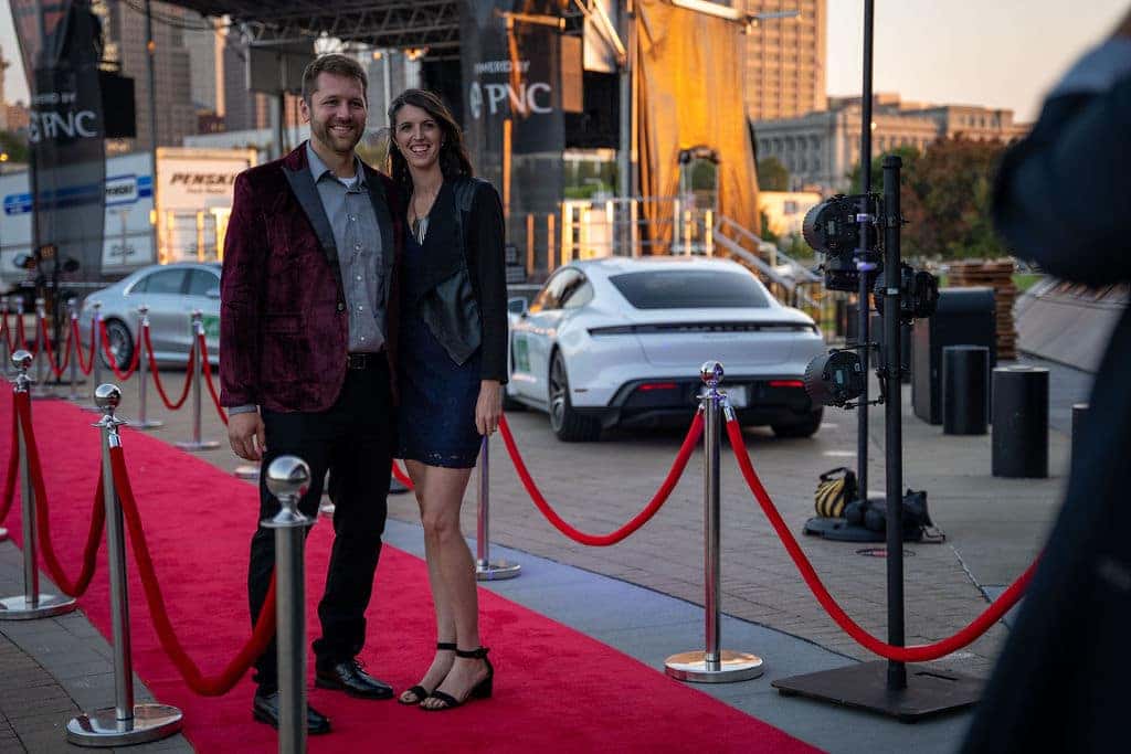 A smiling couple on a red carpet. The man wears a burgundy jacket, and the woman a navy dress. Cars are in the background.