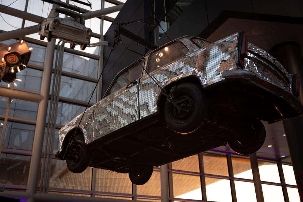 A car covered in glittering silver squares hangs from the ceiling. Another car is above it.