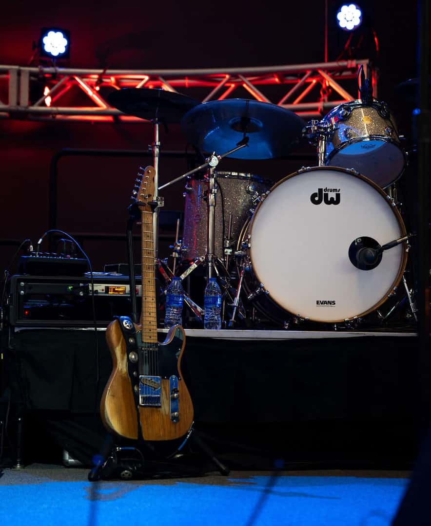A wooden electric guitar on a stand
in front of a drum kit with blue and red stage lights.