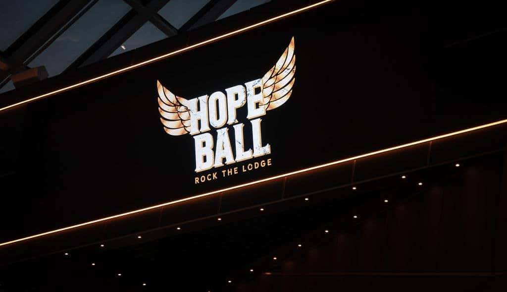 A DJ works mixing equipment on a black table with a glowing HOPE BALL sign above in a dark room.
